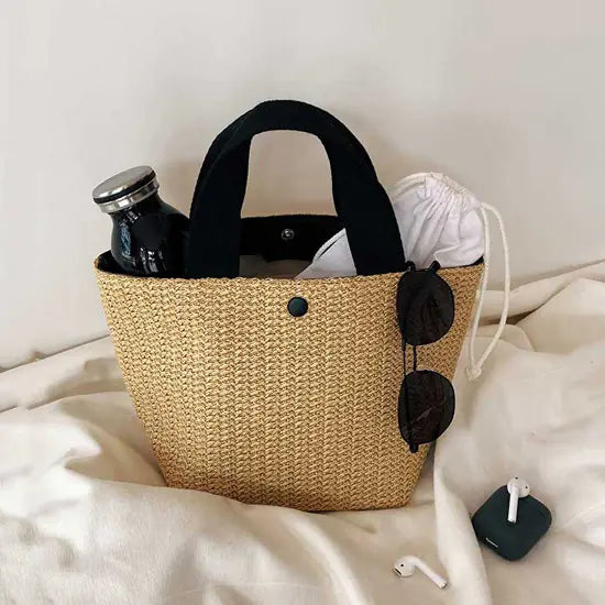 Woven grass straw tote bag with black handles, sunglasses, and a water bottle in beach bucket style.