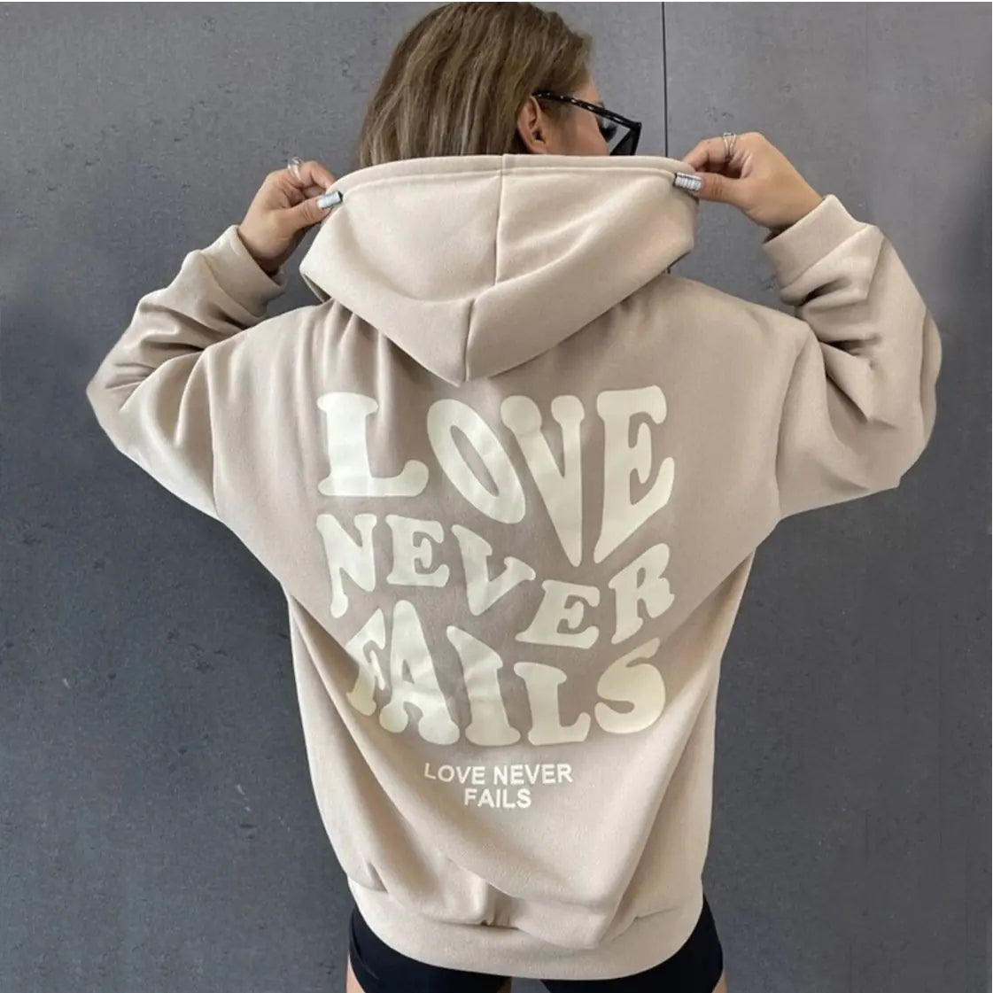Classic beige hoodie with 'LOVE NEVER FAILS' print on the back, perfect for casual styling.