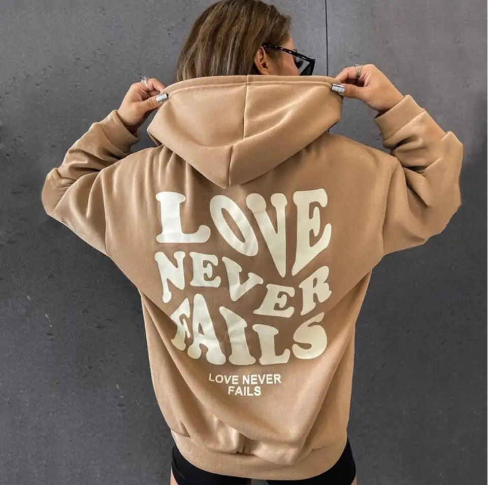 Back view of a woman wearing a stylish beige hoodie with the text 'LOVE NEVER FAILS' printed in bold letters.