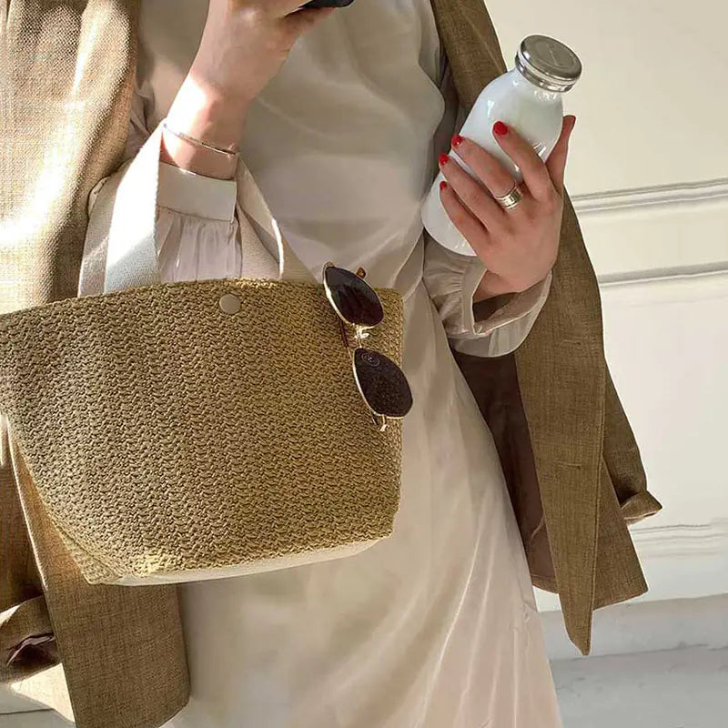 Woven grass straw tote bag in beach bucket style held by a person, featuring sunglasses and a milk bottle.