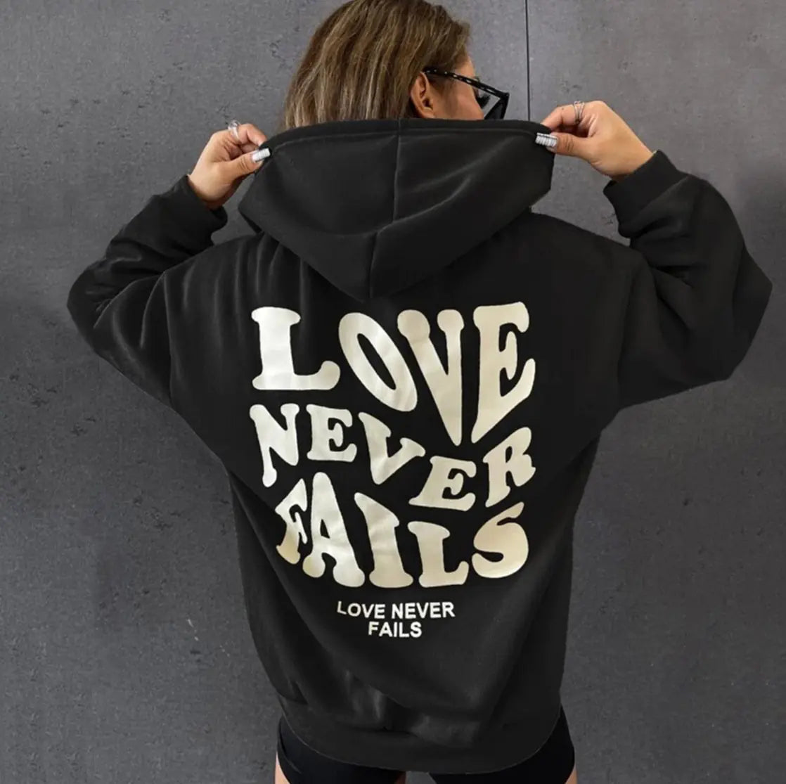 Back view of a woman wearing a black hoodie with the phrase 'Love Never Fails' printed on it, showcasing a stylish streetwear look.