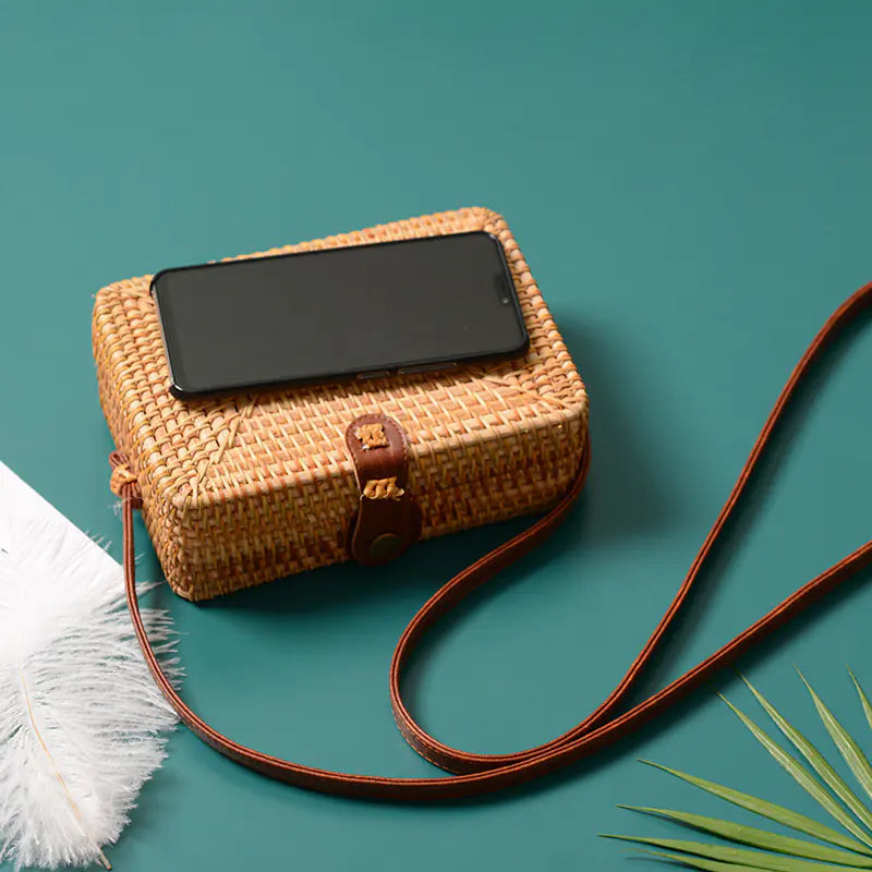 Woven rattan mini bag with leather closure and smartphone on teal background, showcasing retro style and craftsmanship.
