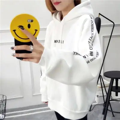 Woman wearing a white hoodie with printed text, holding a smiley face phone case, showcasing casual street style.