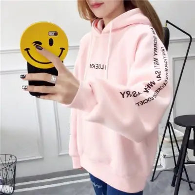 Woman wearing a pink hoodie with a hood and black text design, holding a smiley face phone case.