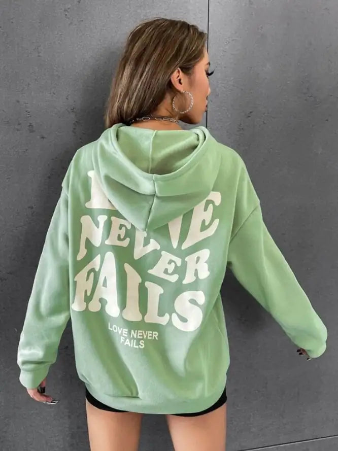 Woman wearing a green hoodie with 'Love Never Fails' printed on the back, showcasing a relaxed fit.