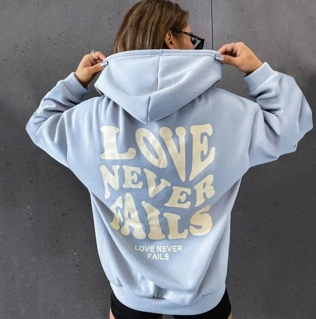 Woman wearing a light blue hoodie with the phrase 'Love Never Fails' printed on the back, standing against a gray wall.