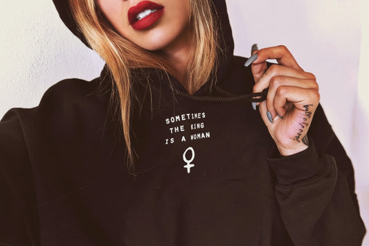 Model wearing SOMETIMES THE KING IS A WOMAN Hoodie in black with bold text and feminine symbol.