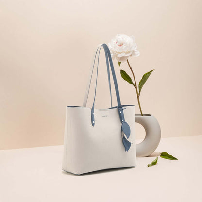 Large capacity leather handbag in white with blue straps, elegant design displayed beside a plant and vase.